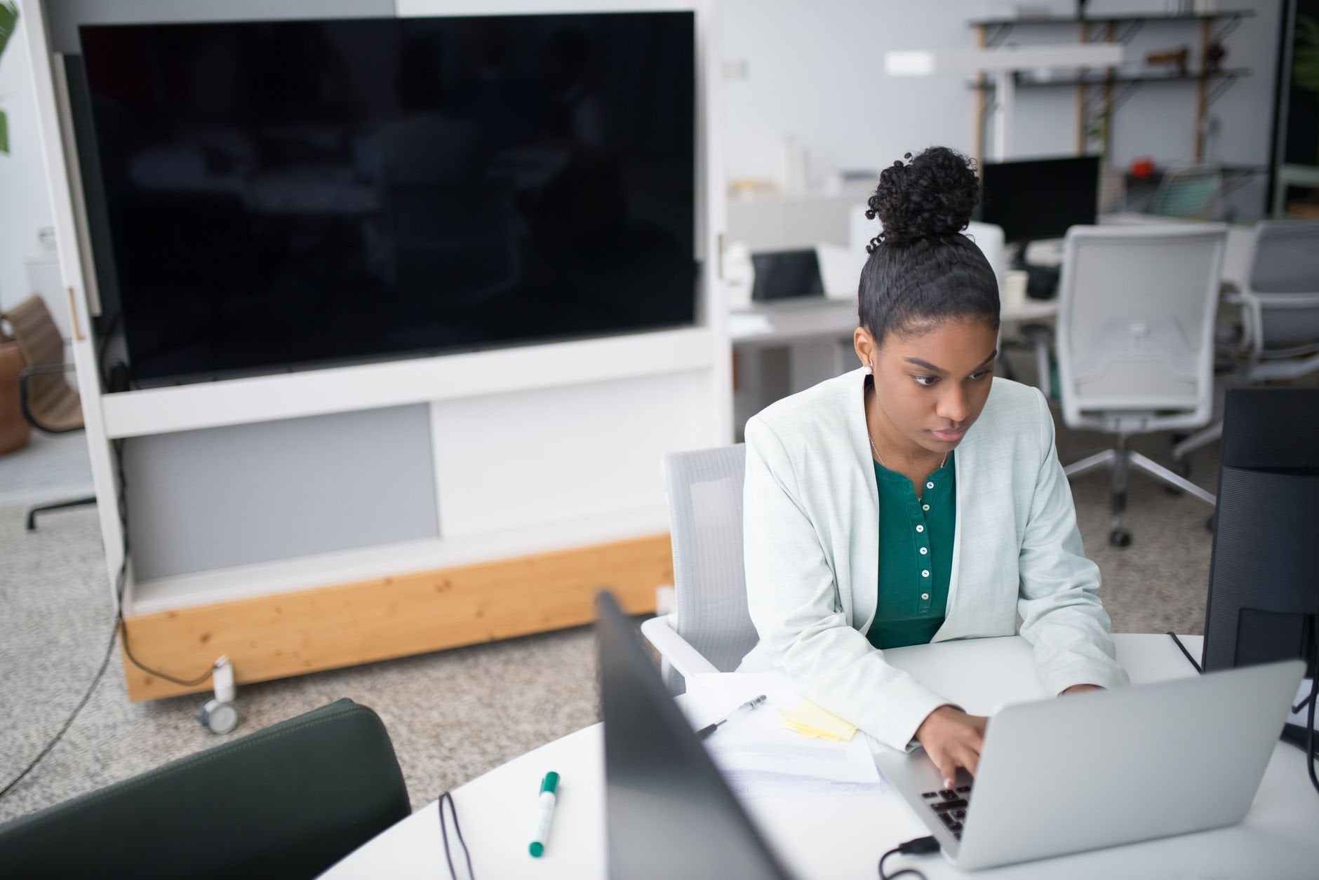 Focused woman using a laptop in a modern office setting, wearing a white blazer, working diligently.